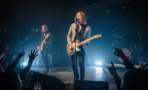 A rock band performs on stage, with lead singer and guitarist in the foreground.