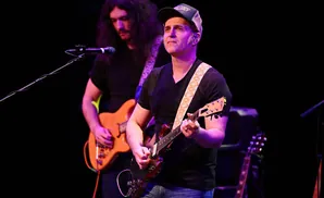 A man in a baseball cap plays a guitar while another man with long hair plays a guitar behind him.