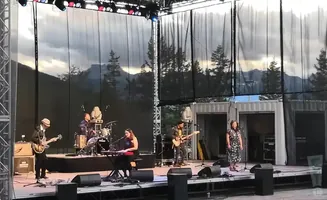 A band performs on an outdoor stage with a mountain and tree backdrop.