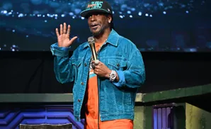Comedian Katt Williams wearing a teal denim jacket and a Miami Dolphins hat on stage.
