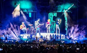 A band performs on a stage with animated characters projected behind them, while a crowd records them on their phones.