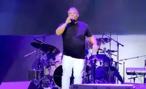 Singer in black shirt and white pants on stage with drums and keyboard.