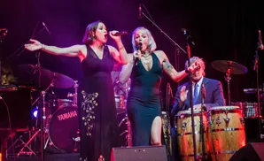 Two women sing into microphones on stage with a band.