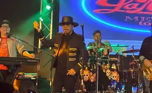 A band performs on stage, with a singer in a black hat and jacket holding a microphone, surrounded by other musicians.