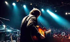 Musician playing an acoustic guitar on stage with blue spotlights and a crowd in the background.