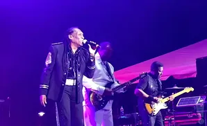 A Black man sings into a microphone on stage, wearing a black jacket with white trim and braids. Two guitarists accompany him.