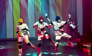 Dancers in colorful outfits and masks perform on a stage with a striped backdrop.
