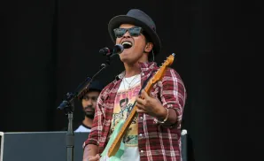 Bruno Mars performs on stage wearing a fedora, sunglasses, and a plaid shirt, playing a light green guitar.