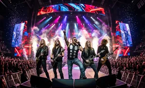 A heavy metal band performs on stage with elaborate lighting and a large screen displaying fiery graphics, while confetti rains down on the audience.