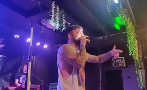 A tattooed man sings into a microphone on a stage lit by purple and green lights, with hanging ivy decorations.