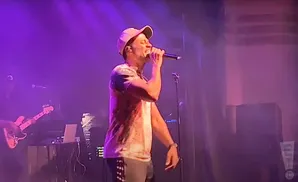 A man in a baseball cap and t-shirt sings into a microphone on a stage with purple lighting.