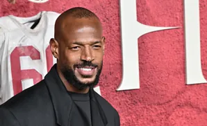 Maricon Wayans smiles in a black suit, with a white jersey bearing the number 6 blurred behind him on a red textured background.