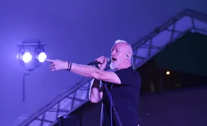 Man with white hair singing and pointing, illuminated by blue stage lights.
