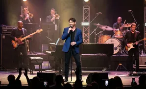 A band performs on stage with a singer in a blue jacket in the foreground.