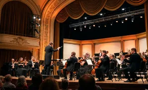 An orchestra conductor leads a performance on stage, with musicians playing various instruments.
