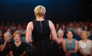 A woman in a sparkly black dress faces away from the camera, addressing an audience.
