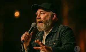 A man with a beard wearing a black baseball cap and denim jacket speaks into a microphone.