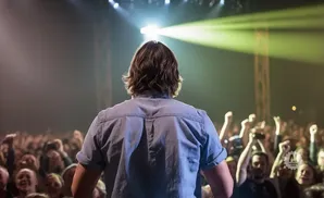 A person in a blue shirt faces a cheering crowd with their back to the camera, illuminated by stage lights.