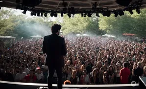 A man in a suit stands on stage facing a large, enthusiastic crowd at an outdoor concert.