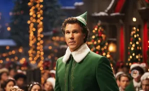 Will Ferrell as Buddy the Elf in a green elf costume with a white fur collar, standing in front of Christmas trees and lights.