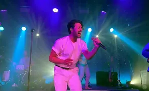 A male singer in white performs on stage, bathed in blue and purple lights.