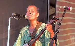 A woman with her hair pulled back sings into a microphone while playing a banjo.