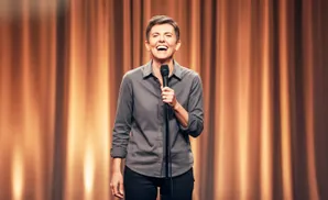 A woman with short hair laughs while holding a microphone on a stage with orange curtains.