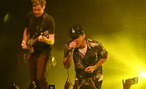 A musician with a LA Dodgers cap sings into a microphone on a stage lit by yellow lights.