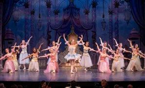 Ballet dancers in tutus and tiaras perform on a stage with a regal backdrop.