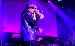 Rapper performing on stage in a bucket hat, sunglasses, and a blue vest over a black hoodie.