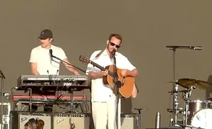 Musicians perform on stage; one plays guitar and sings, another plays a keyboard.