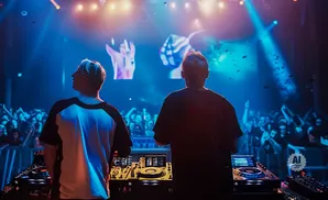 Two DJs perform on stage in front of a cheering crowd, illuminated by blue and pink stage lights.