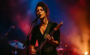 A female guitarist sings into a microphone on a dimly lit stage with red and blue lights.