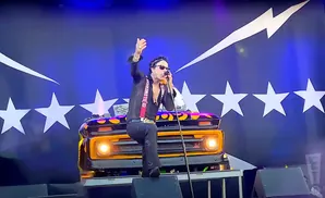 A man in sunglasses and a black shirt with red suspenders sings into a microphone on stage in front of a blue backdrop with white stars.