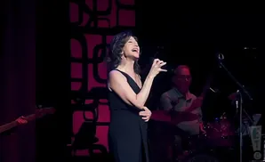 Singer Amy Grant performs in a black dress on stage, with a drummer and bassist visible.