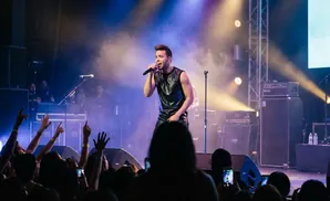 A male singer in a black leather outfit performs on stage, holding a microphone, with a crowd in the foreground.