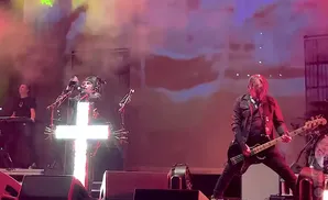 A band performs on stage with bright lights, a lit cross, and smoke effects.