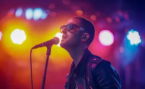 A singer wearing sunglasses and a leather jacket performs into a microphone on a brightly lit stage.