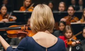 A violinist in a blue dress plays in an orchestra, with other musicians blurred in the background.