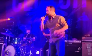 A singer with a beard performs on stage with a drummer in the background.