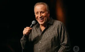 A man with a wide smile holds a microphone and wears a striped shirt.