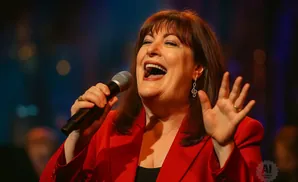 A woman in a red jacket sings into a microphone, her mouth open in song.