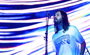 A man with long hair and a beard sings into a microphone on a stage with a colorful, striped background.