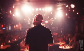 A bald man with glasses on a stage faces a blurred audience under bright stage lights.