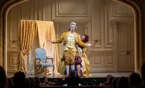 Opera singers on stage in elaborate costumes, with an orchestra in the foreground.