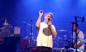 A man in a white t-shirt sings into a microphone on a stage with a blue backdrop.
