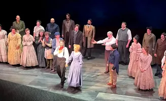 A cast of actors in historical costumes stand on a stage, some holding hands and smiling.