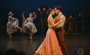 Ballet dancers in period costumes perform on stage. A couple in the foreground dances intimately, while others move in the background.