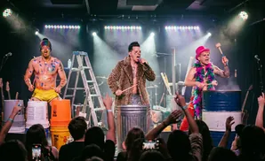 Three musicians perform on stage with colorful outfits and body paint, playing drums made of buckets and barrels.
