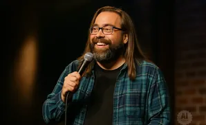 A man with glasses, a beard, and long hair laughs while holding a microphone, wearing a blue plaid shirt.